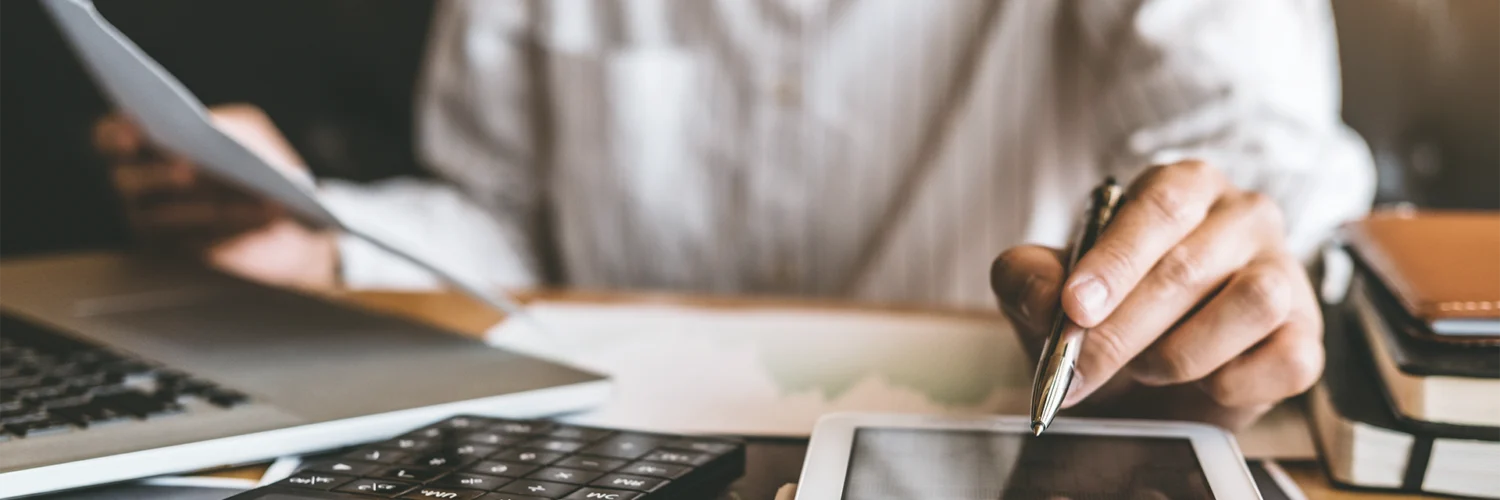 Pessoa trabalhando com documentos, tablet, laptop e calculadora sobre a mesa em ambiente de escritório.