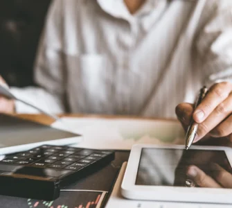 Pessoa trabalhando com documentos, tablet, laptop e calculadora sobre a mesa em ambiente de escritório.