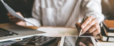Pessoa trabalhando com documentos, tablet, laptop e calculadora sobre a mesa em ambiente de escritório.
