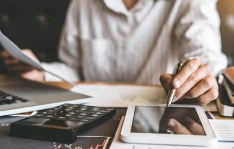 Pessoa trabalhando com documentos, tablet, laptop e calculadora sobre a mesa em ambiente de escritório.