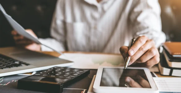 Pessoa trabalhando com documentos, tablet, laptop e calculadora sobre a mesa em ambiente de escritório.