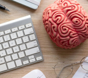 Mesa de trabalho com teclado, óculos, questionário, cérebro de plástico e itens de papelaria.