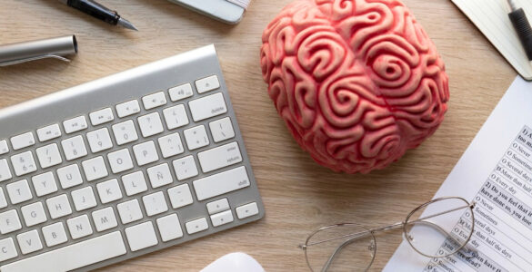 Mesa de trabalho com teclado, óculos, questionário, cérebro de plástico e itens de papelaria.