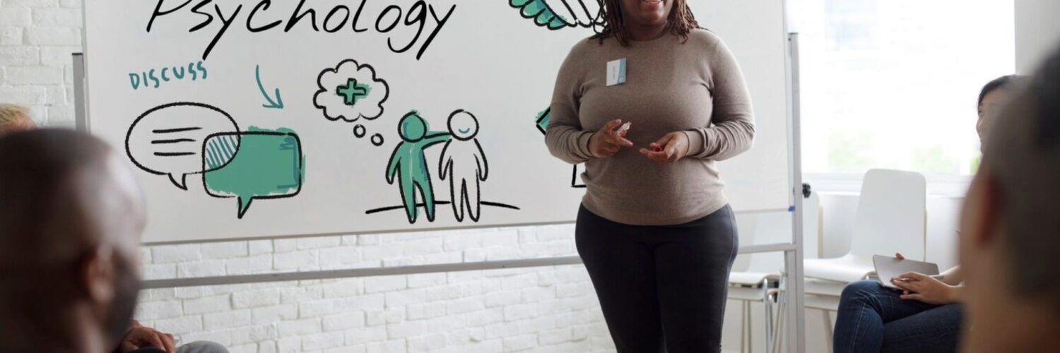 Palestrante apresenta conteúdo sobre Psicologia em sala com grupo e quadro ilustrado ao fundo.