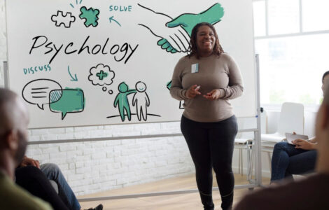 Palestrante apresenta conteúdo sobre Psicologia em sala com grupo e quadro ilustrado ao fundo.