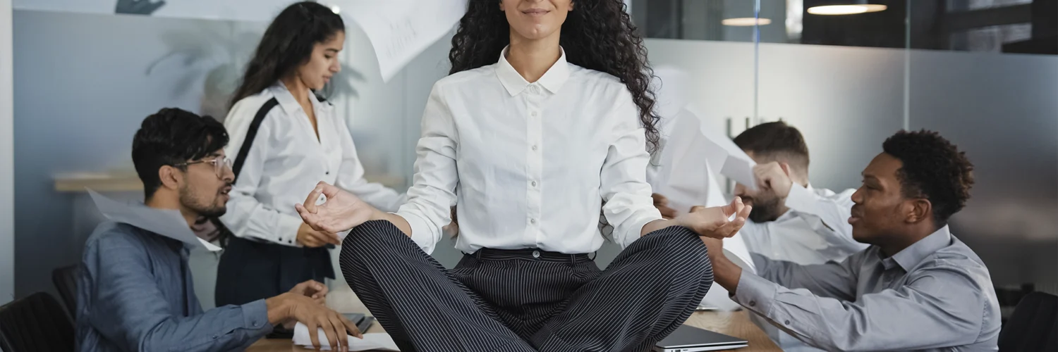 Mulher meditando sobre a mesa em meio ao caos no escritório, simbolizando calma no estresse do trabalho.