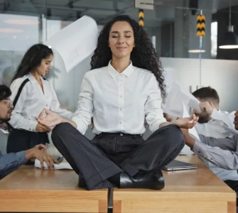 Mulher meditando sobre a mesa em meio ao caos no escritório, simbolizando calma no estresse do trabalho.