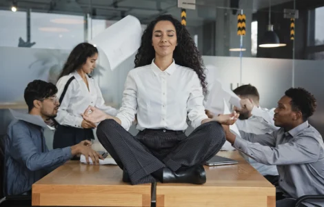 Mulher meditando sobre a mesa em meio ao caos no escritório, simbolizando calma no estresse do trabalho.