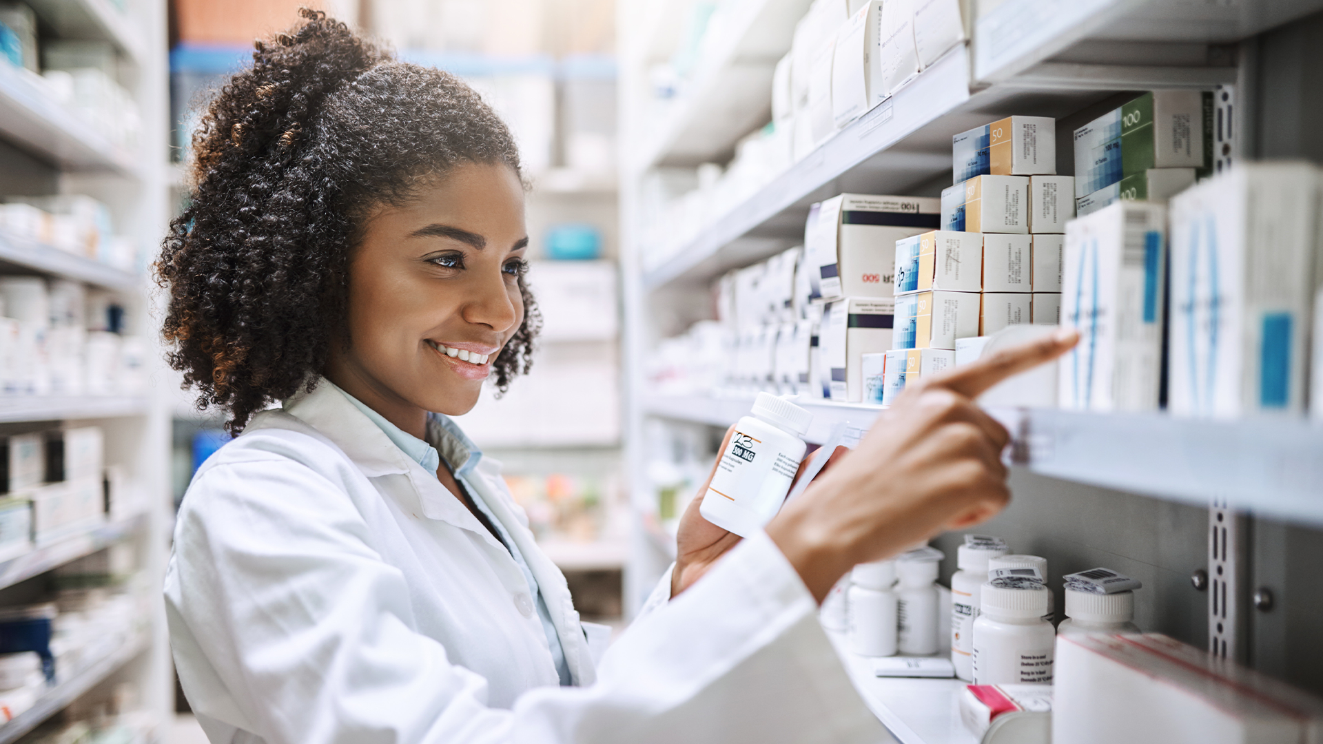 Mulher de jaleco sorrindo enquanto organiza medicamentos em prateleira de farmácia.
