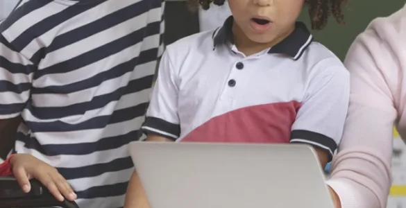Professora e crianças, incluindo aluna em cadeira de rodas, interagem com laptop em sala de aula inclusiva.