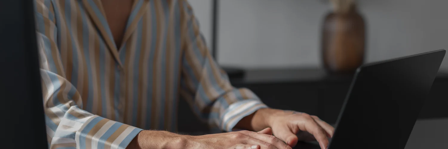 Mulher digitando em laptop em ambiente de escritório, vestindo camisa listrada e colar de pérolas.