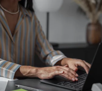 Mulher digitando em laptop em ambiente de escritório, vestindo camisa listrada e colar de pérolas.