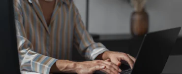 Mulher digitando em laptop em ambiente de escritório, vestindo camisa listrada e colar de pérolas.