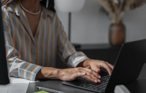 Mulher digitando em laptop em ambiente de escritório, vestindo camisa listrada e colar de pérolas.