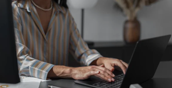 Mulher digitando em laptop em ambiente de escritório, vestindo camisa listrada e colar de pérolas.