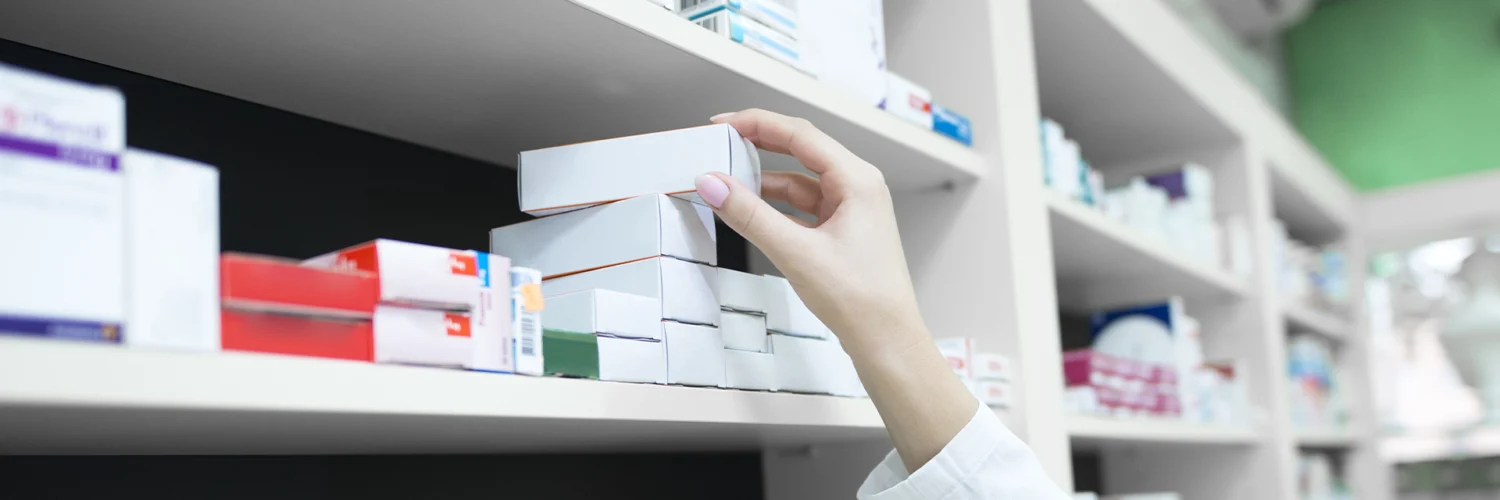 Pessoa com jaleco pegando caixa de medicamento em prateleira de farmácia organizada com vários remédios.