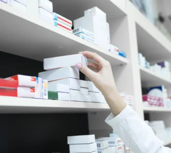 Pessoa com jaleco pegando caixa de medicamento em prateleira de farmácia organizada com vários remédios.
