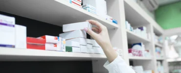Pessoa com jaleco pegando caixa de medicamento em prateleira de farmácia organizada com vários remédios.