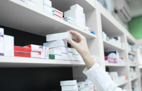 Pessoa com jaleco pegando caixa de medicamento em prateleira de farmácia organizada com vários remédios.