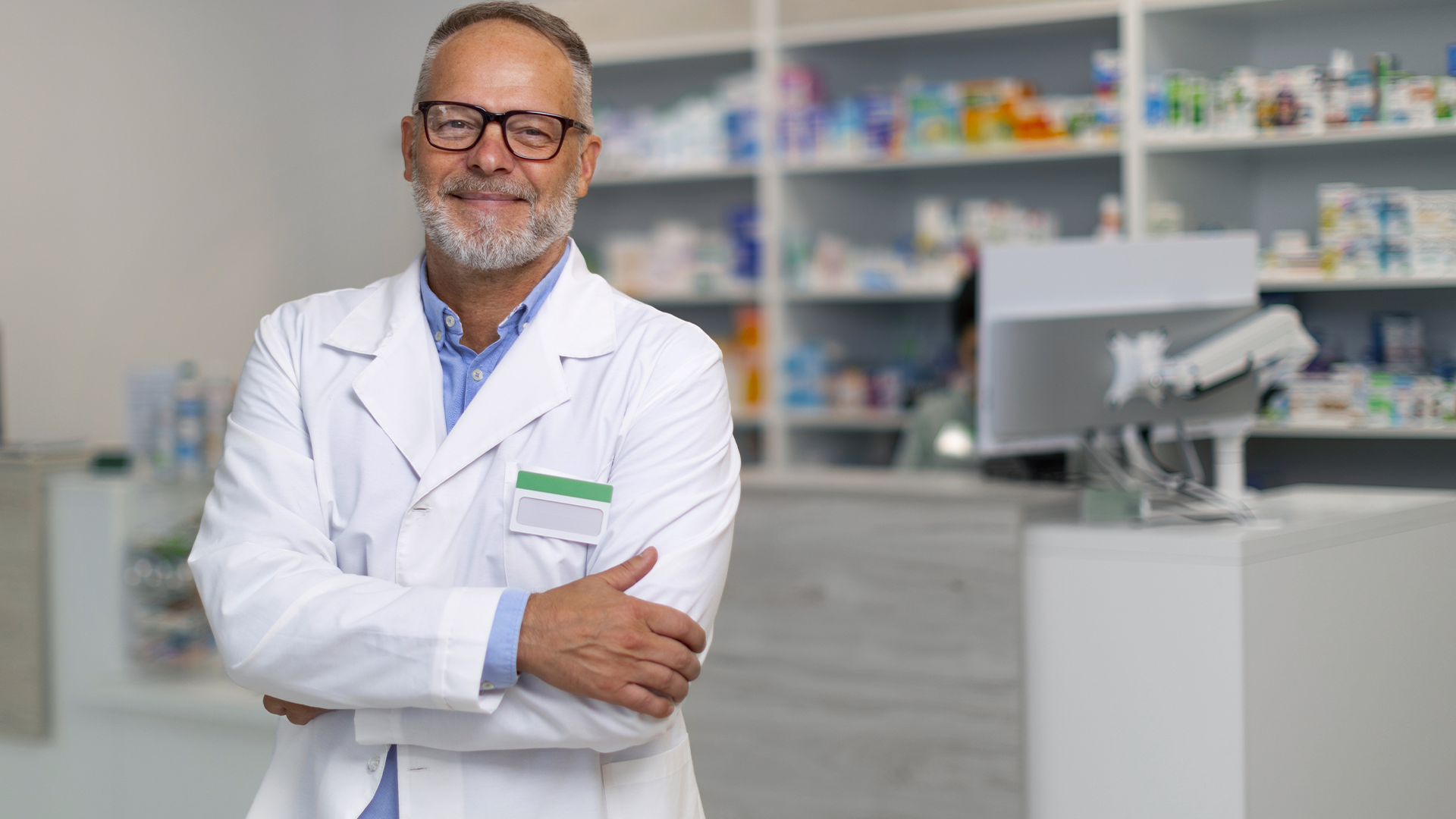 Farmacêutico sorridente de jaleco branco em frente ao balcão de farmácia, com prateleiras de medicamentos ao fundo.