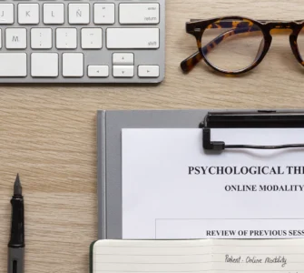 Mesa com teclado, óculos, caneta e ficha de terapia psicológica online com anotações de sessões anteriores.