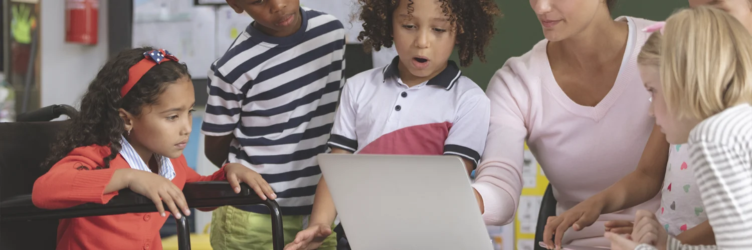 Professora e crianças, incluindo aluna em cadeira de rodas, interagem com laptop em sala de aula inclusiva.
