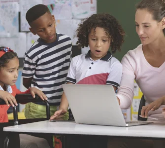 Professora e crianças, incluindo aluna em cadeira de rodas, interagem com laptop em sala de aula inclusiva.
