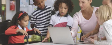 Professora e crianças, incluindo aluna em cadeira de rodas, interagem com laptop em sala de aula inclusiva.