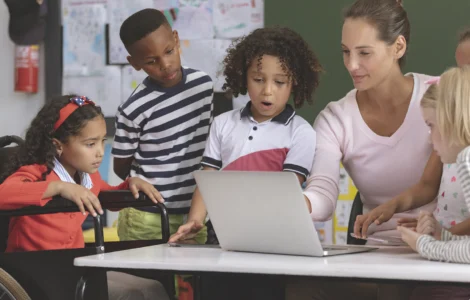 Professora e crianças, incluindo aluna em cadeira de rodas, interagem com laptop em sala de aula inclusiva.