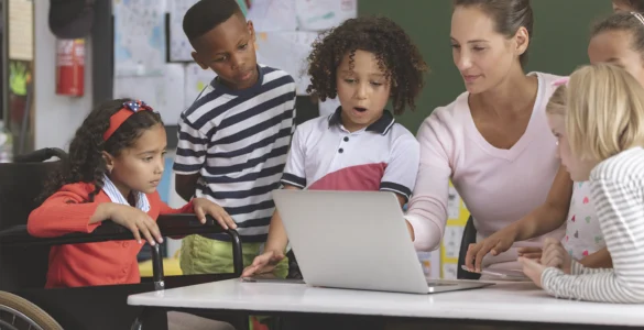Professora e crianças, incluindo aluna em cadeira de rodas, interagem com laptop em sala de aula inclusiva.