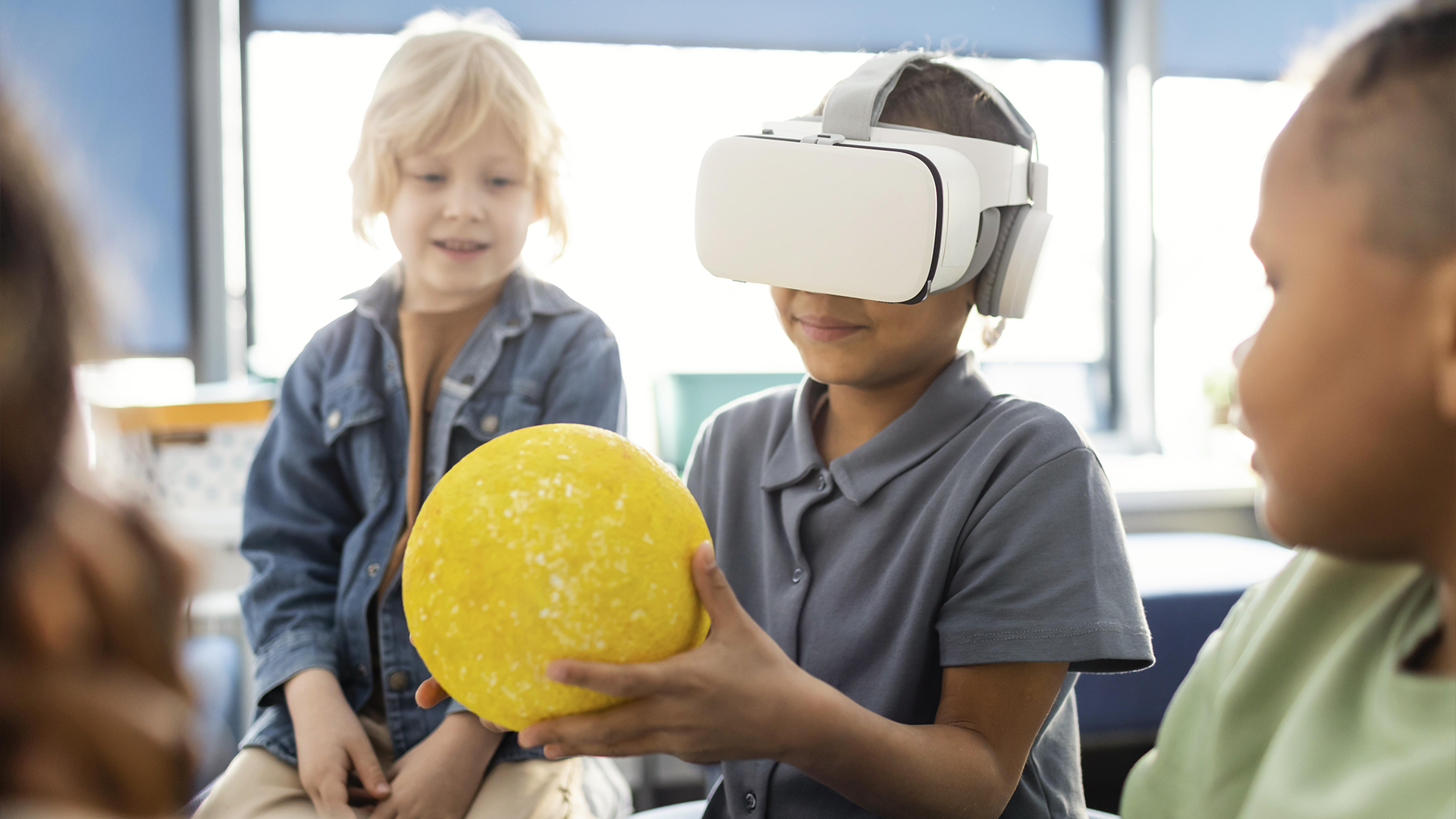 Criança com óculos de realidade virtual segura modelo do Sol enquanto colegas observam em sala de aula.