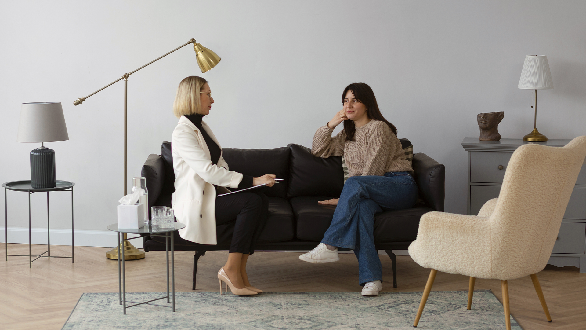Duas mulheres sentadas em uma sala: uma terapeuta loira com blazer branco e uma paciente de suéter bege e jeans.