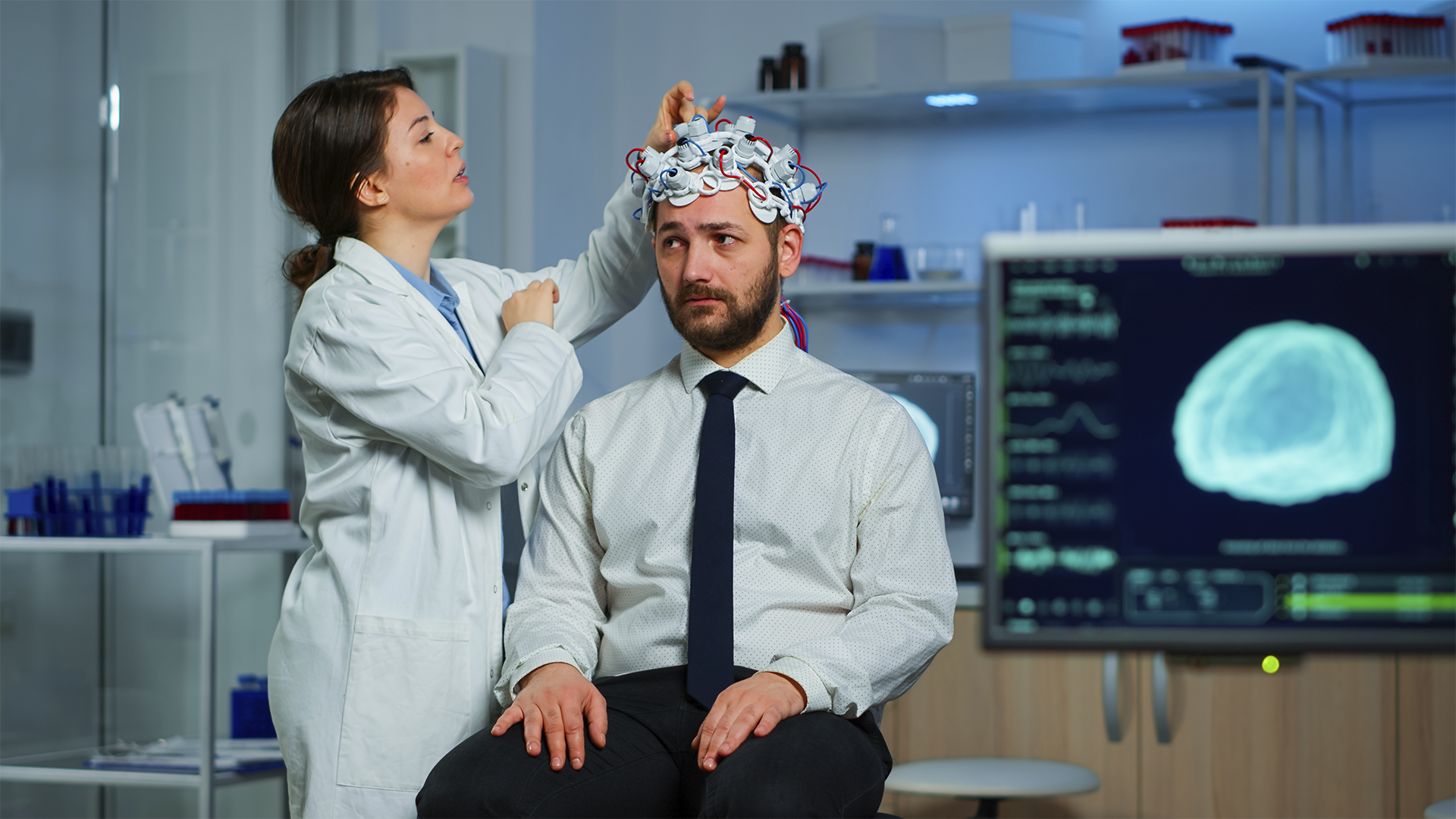 Uma cientista coloca eletrodos em um homem para um eletroencefalograma (EEG), com um monitor mostrando um cérebro ao fundo.