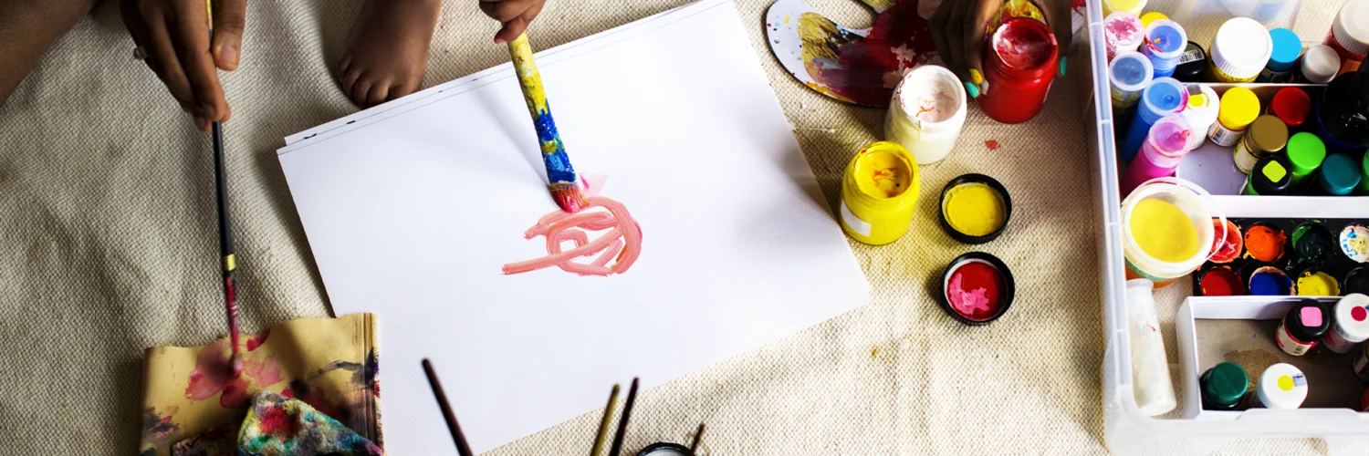 Crianças e adultos pintando com tintas coloridas sobre papel, rodeados por materiais de arte diversos.