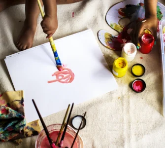 Crianças e adultos pintando com tintas coloridas sobre papel, rodeados por materiais de arte diversos.