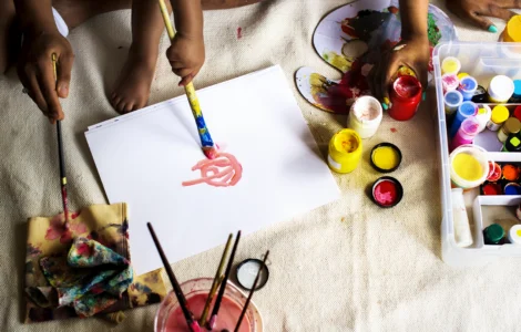Crianças e adultos pintando com tintas coloridas sobre papel, rodeados por materiais de arte diversos.