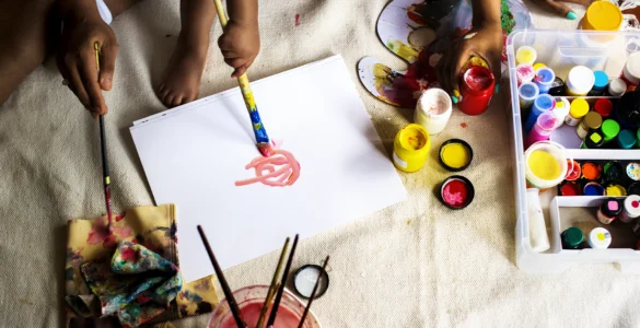 Crianças e adultos pintando com tintas coloridas sobre papel, rodeados por materiais de arte diversos.
