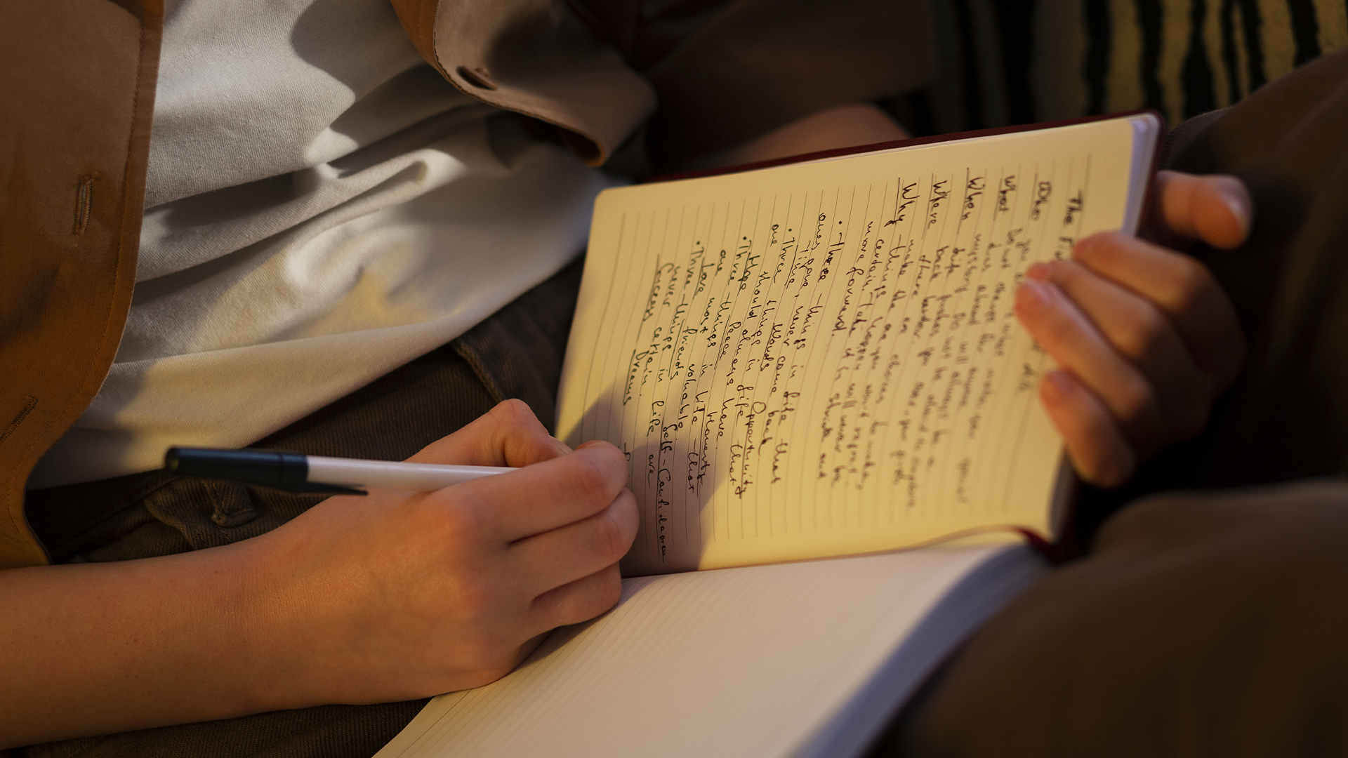 Corte aproximado de uma pessoa escrevendo notas em um caderno, segurando uma caneta, com luz indireta iluminando as páginas.