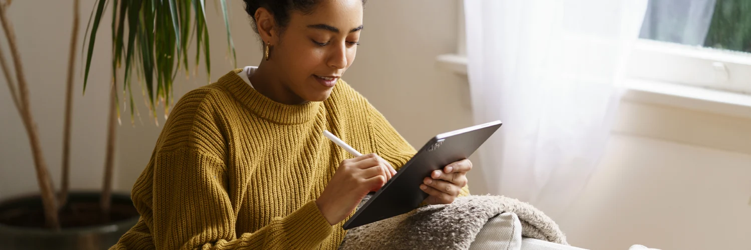 Mulher sorridente com cabelo cacheado, vestindo um suéter mostarda, sentada no sofá usando um tablet com caneta.