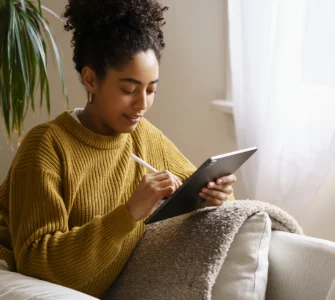 Mulher sorridente com cabelo cacheado, vestindo um suéter mostarda, sentada no sofá usando um tablet com caneta.