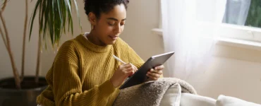 Mulher sorridente com cabelo cacheado, vestindo um suéter mostarda, sentada no sofá usando um tablet com caneta.