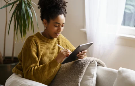 Mulher sorridente com cabelo cacheado, vestindo um suéter mostarda, sentada no sofá usando um tablet com caneta.
