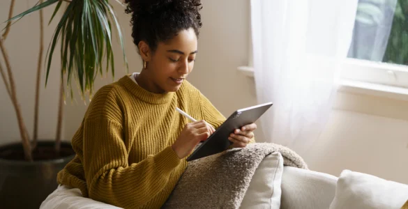 Mulher sorridente com cabelo cacheado, vestindo um suéter mostarda, sentada no sofá usando um tablet com caneta.