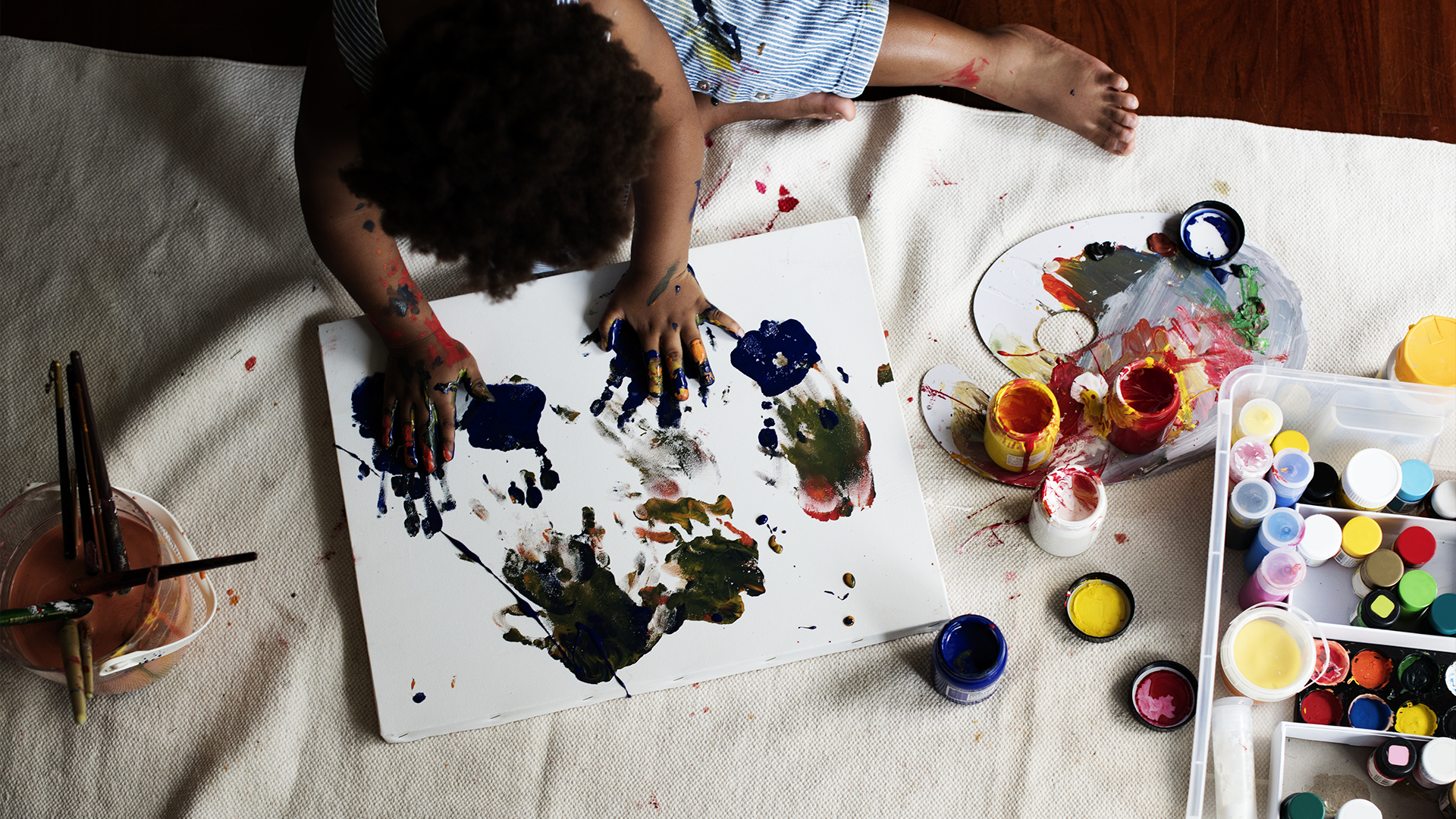 Criança pinta com as mãos em tela no chão, rodeada por tintas coloridas, pincéis e paleta de pintura.