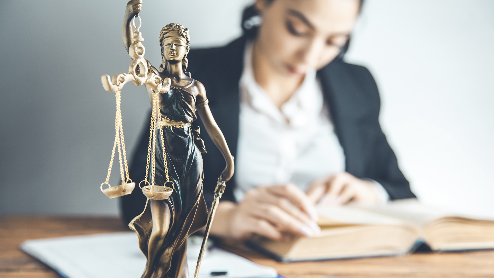Estátua da justiça em primeiro plano, com uma mulher estudando um livro jurídico desfocada ao fundo.