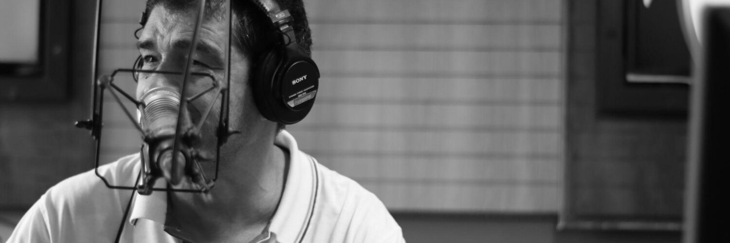 Homem com fones de ouvido fala ao microfone em estúdio de rádio, em imagem em preto e branco.