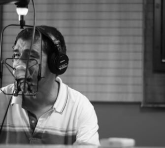 Homem com fones de ouvido fala ao microfone em estúdio de rádio, em imagem em preto e branco.