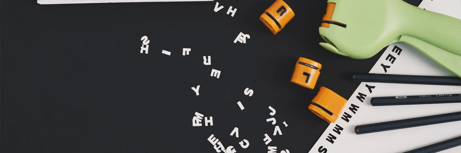 Uma mesa preta com letras e números espalhados, dois blocos laranja, um livro e lápis, sugerindo estudo.