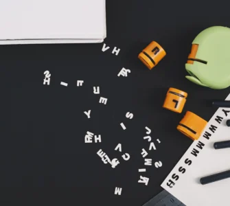 Uma mesa preta com letras e números espalhados, dois blocos laranja, um livro e lápis, sugerindo estudo.