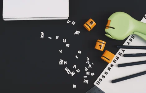 Uma mesa preta com letras e números espalhados, dois blocos laranja, um livro e lápis, sugerindo estudo.